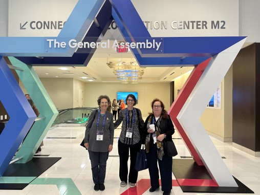 Three women stand together in front of a large, colorful star-shaped installation that reads "The General Assembly." They are inside a convention center.