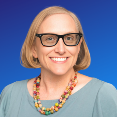 A woman with short blonde hair and black glasses smiles at the camera, wearing a light blue top and a colorful beaded necklace, against a blue background.